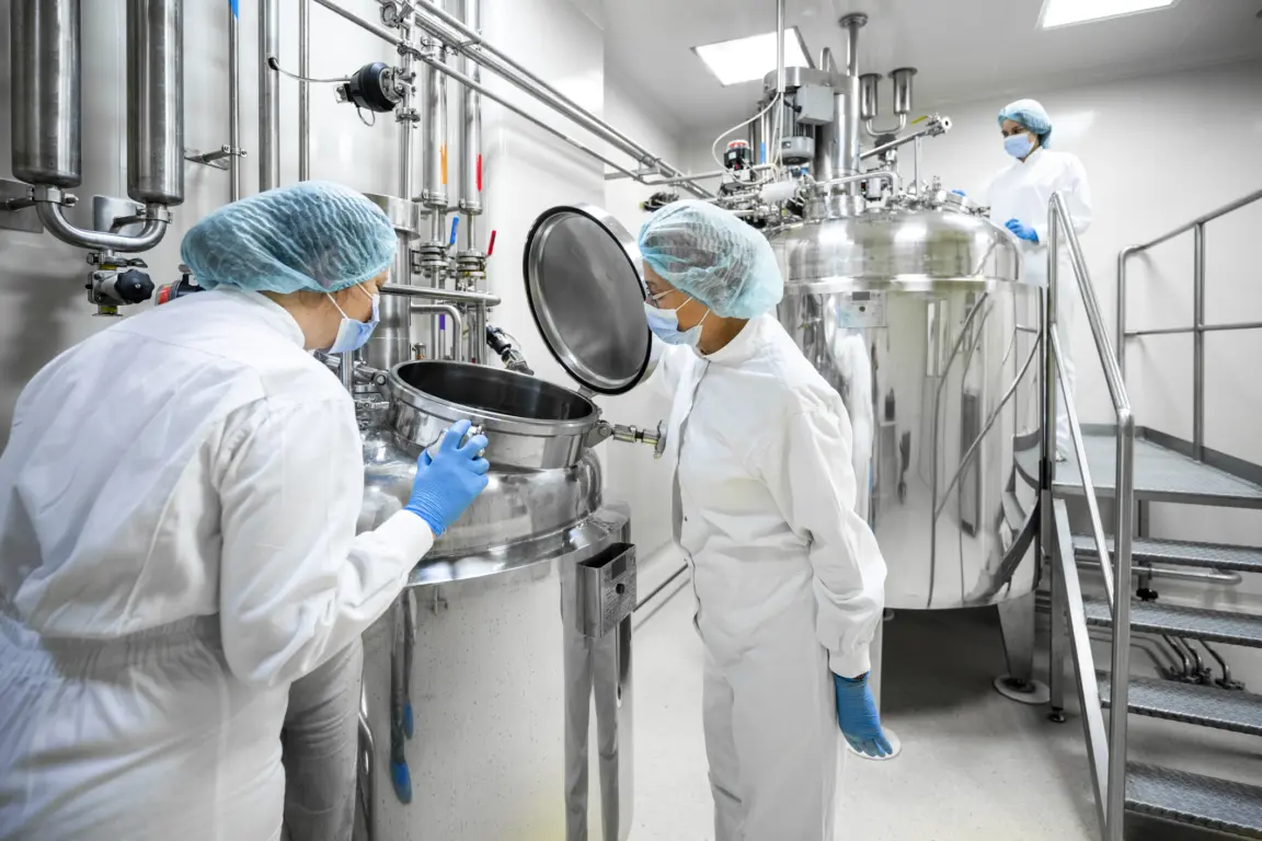 Three female workers in pharmaceutical industry wearing protective blue gloves, mask and cap and white suit seen standing by the machine that is the part of the medicaments production during the working hours in a pharmaceutical manufacturing.