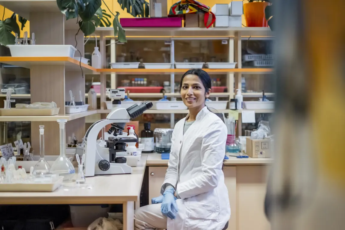 An Indian female researcher, wearing a white lab coat and gloves, smiles while working in a modern university laboratory equipped with a microscope and scientific tools. The vibrant lab environment features organized shelves, plants, and scientific equipment.