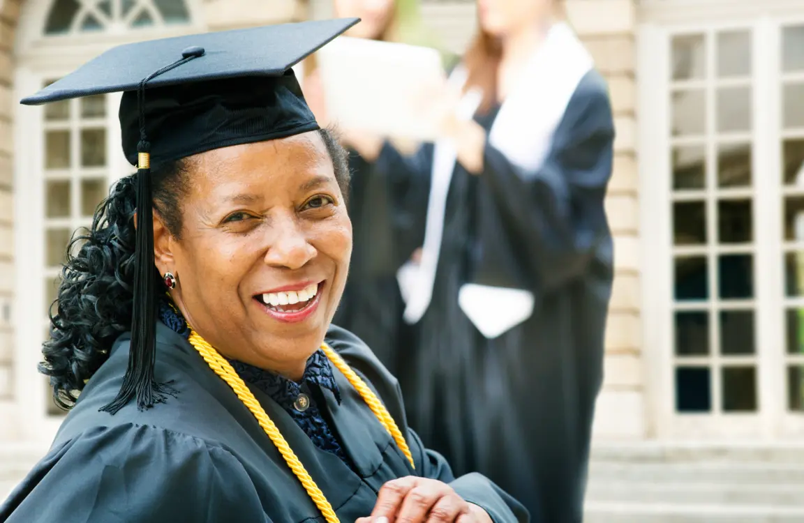 Mature African American woman on graduation day, turning to the camera with a smile. Younger graduates are coming down the stairs behind her.
